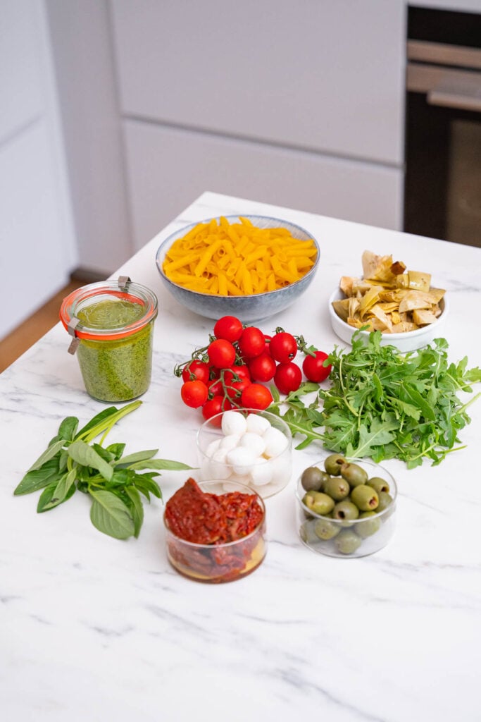 Un comptoir de cuisine avec des bols de poivron jaune tranché, de roquette, de tomates cerises, de boules de mozzarella, d'olives, de tomates séchées au soleil, de pesto, de feuilles de basilic et de croûtons.