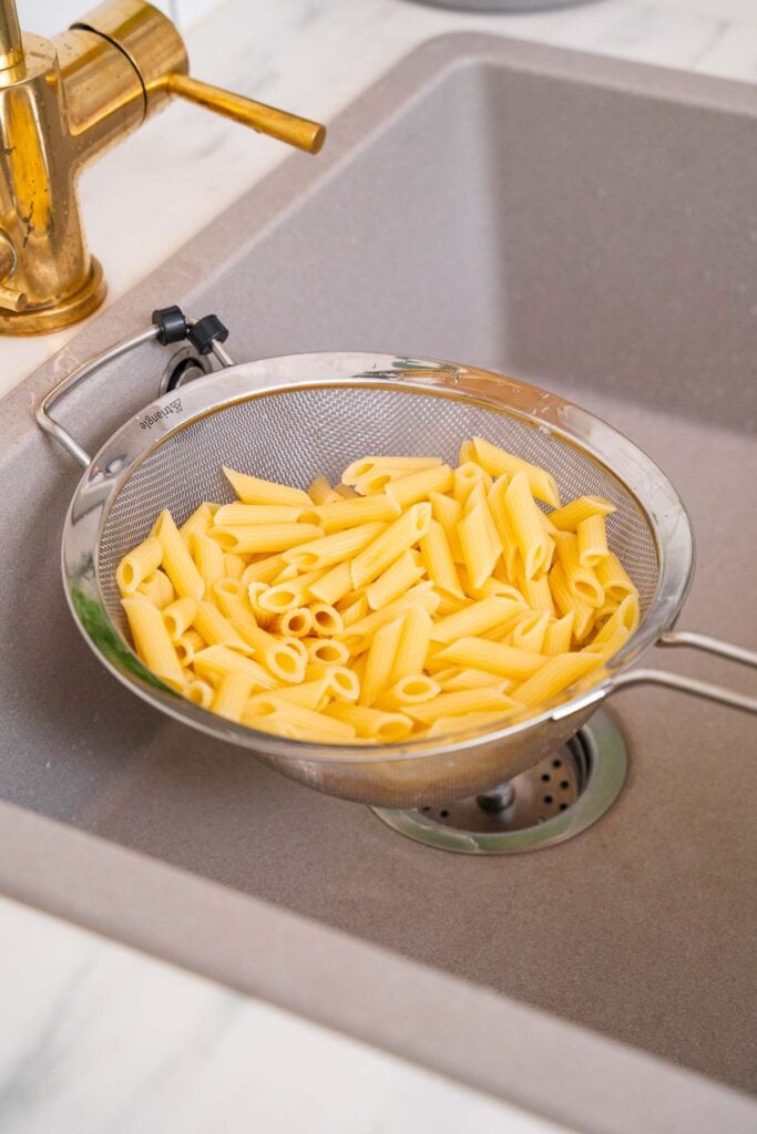 Une passoire remplie de pâtes penne cuites est placée dans un évier de cuisine beige à côté d'un robinet doré.