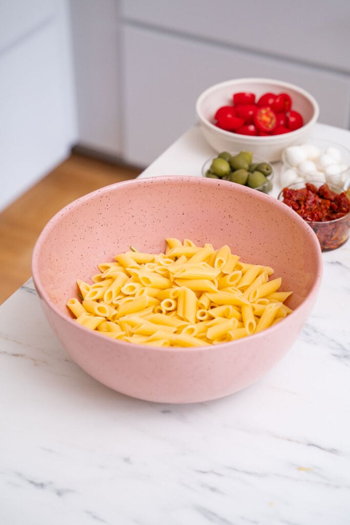 Un bol rose de pâtes crues est posé sur un plan de travail en marbre blanc. À l'arrière-plan, on trouve de petits bols contenant des tomates cerises, des olives vertes, des boules de mozzarella et des tomates séchées au soleil.