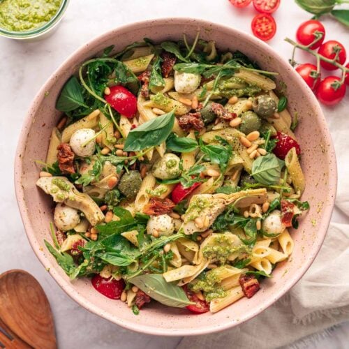 Un bol de salade de pâtes penne avec des boules de mozzarella, des tomates cerises, du basilic frais et du pesto, garni de pignons de pin. Deux cuillères en bois et des ingrédients supplémentaires sont posés sur la surface en marbre à côté du bol.