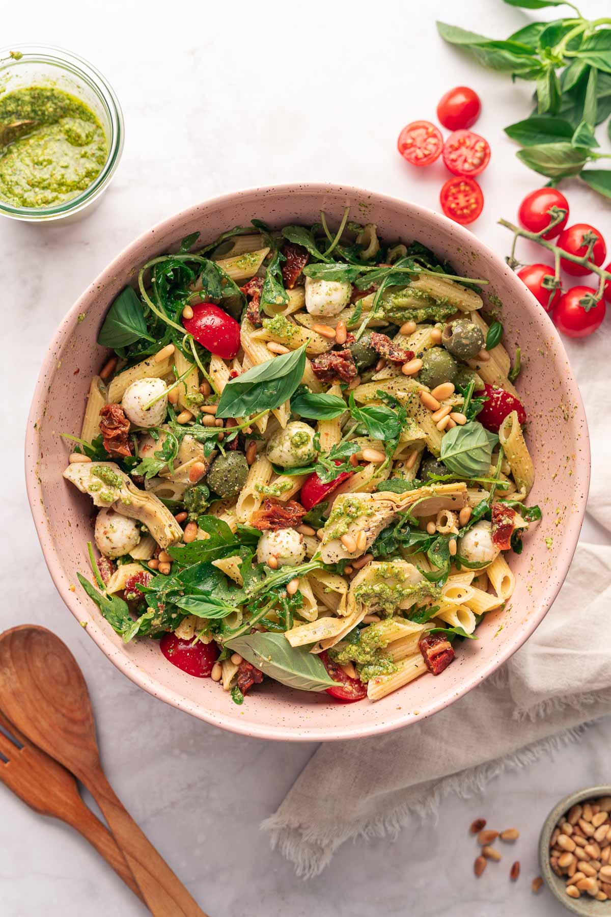 A bowl of pasta salad with cherry tomatoes, mozzarella balls, basil, pine nuts, and arugula, accompanied by wooden utensils and a jar of pesto on the side.