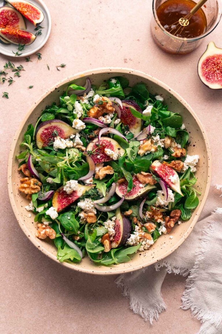 Eine Schüssel mit gemischtem Blattsalat mit in Scheiben geschnittenen Feigen, roten Zwiebeln, zerbröckeltem Ziegenkäse und Walnüssen, garniert mit gemahlenem Pfeffer.
Ein kleiner Glasbehälter mit Dressing steht in der Nähe, ebenso wie eine in Scheiben geschnittene Feige. 