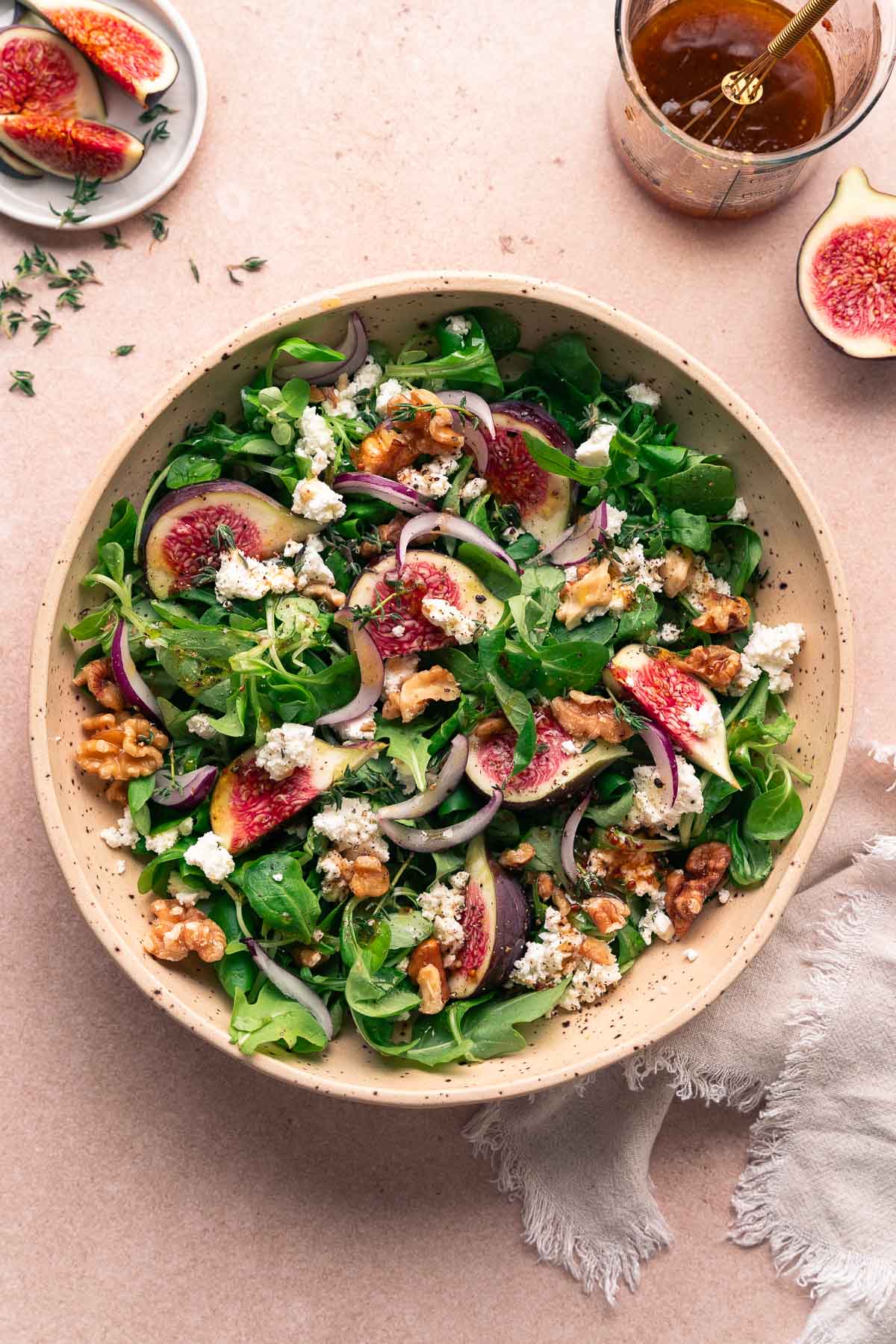 Un bol de salade verte mélangée avec des figues tranchées, des oignons rouges, du chèvre émietté et des noix, agrémenté de poivre moulu. Un petit récipient en verre contenant de la vinaigrette se trouve à proximité, ainsi qu'une figue tranchée.