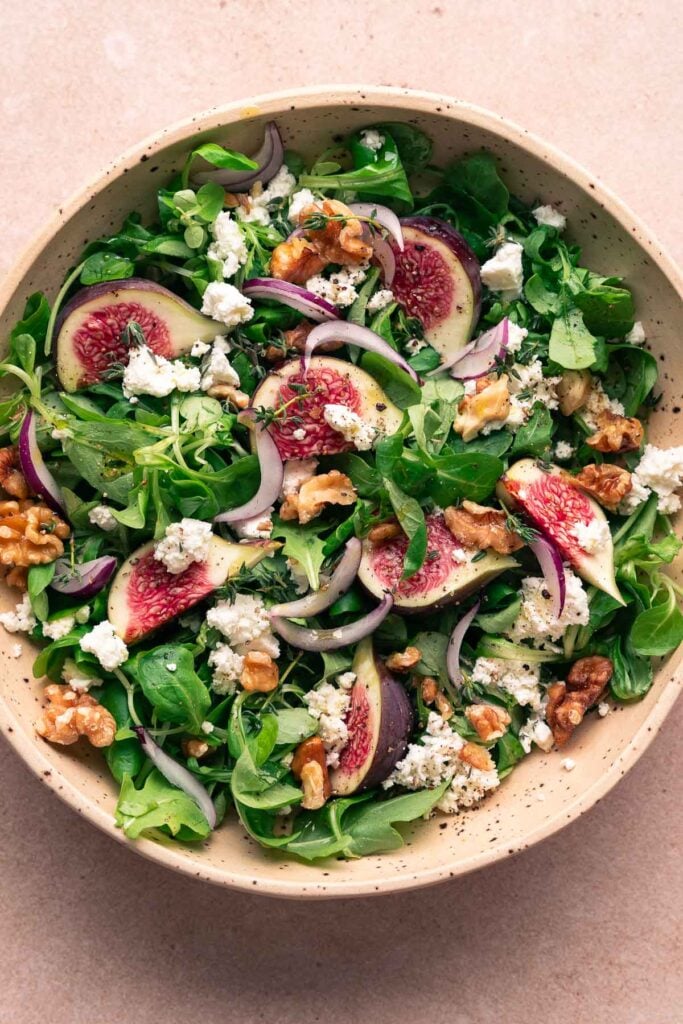 Un bol de salade verte avec des figues fraîches, du fromage feta émietté, des noix, des tranches d'oignon rouge et des herbes.