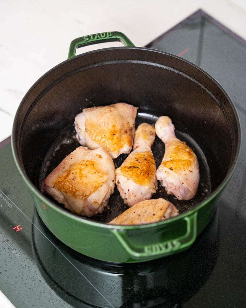 Five pieces of golden chicken cook in a green Staub cast-iron pot on an induction stove.