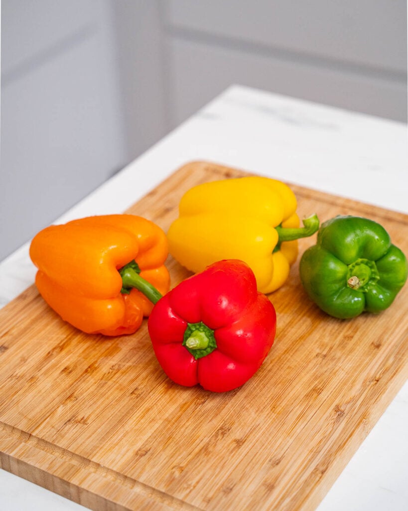 Quatre poivrons, un orange, un jaune, un vert et un rouge, sont placés sur une planche à découper en bois sur un plan de travail blanc.