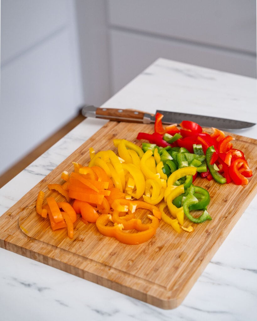 A wooden cutting board on a marble countertop holds chopped red, green, yellow and orange peppers with a knife beside them.