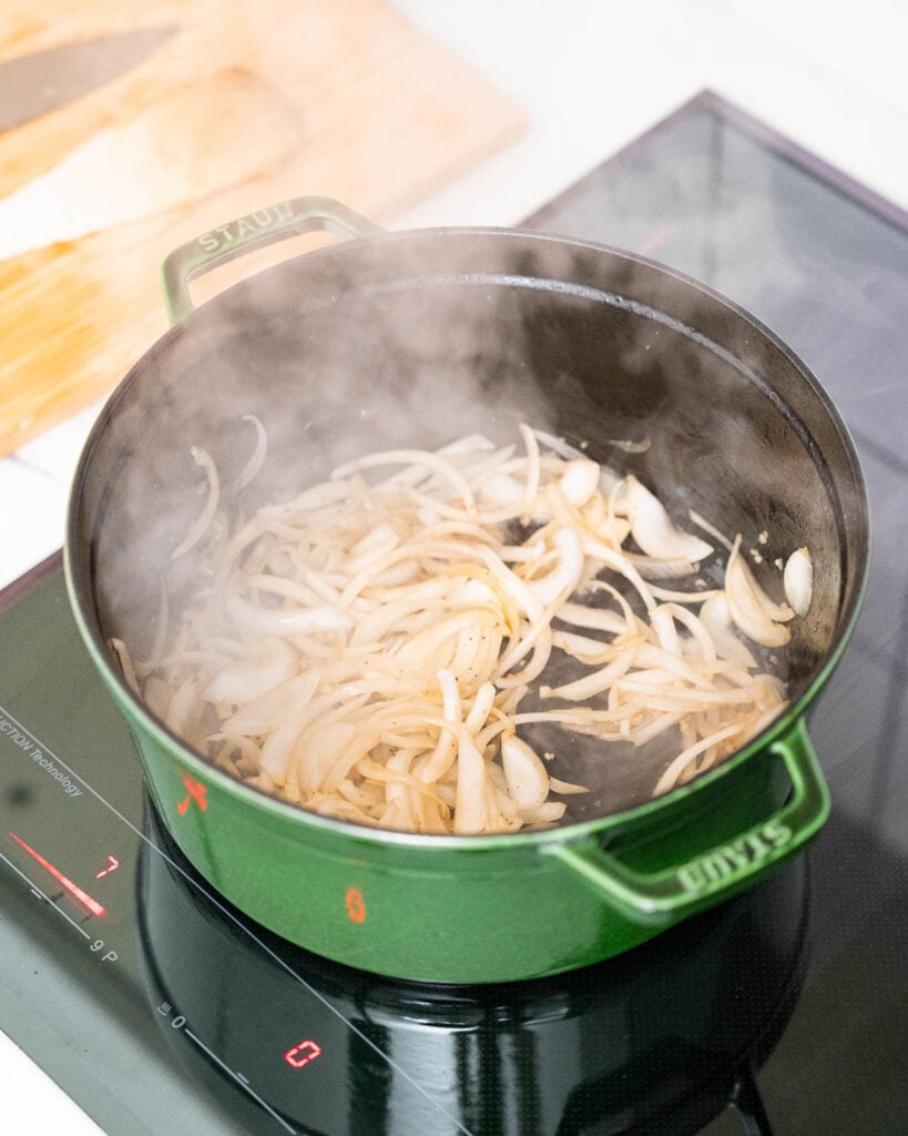 Une casserole verte sur une cuisinière contient des oignons finement tranchés qui sont sautés, avec de la vapeur s'élevant de la casserole.