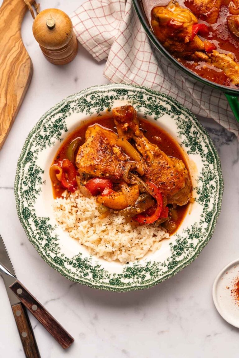 A plate of rice and chicken basquaise with peppers.
A saucepan containing the same dish is partially visible in the background, along with a wooden cutting board, knife and pepper mill. 