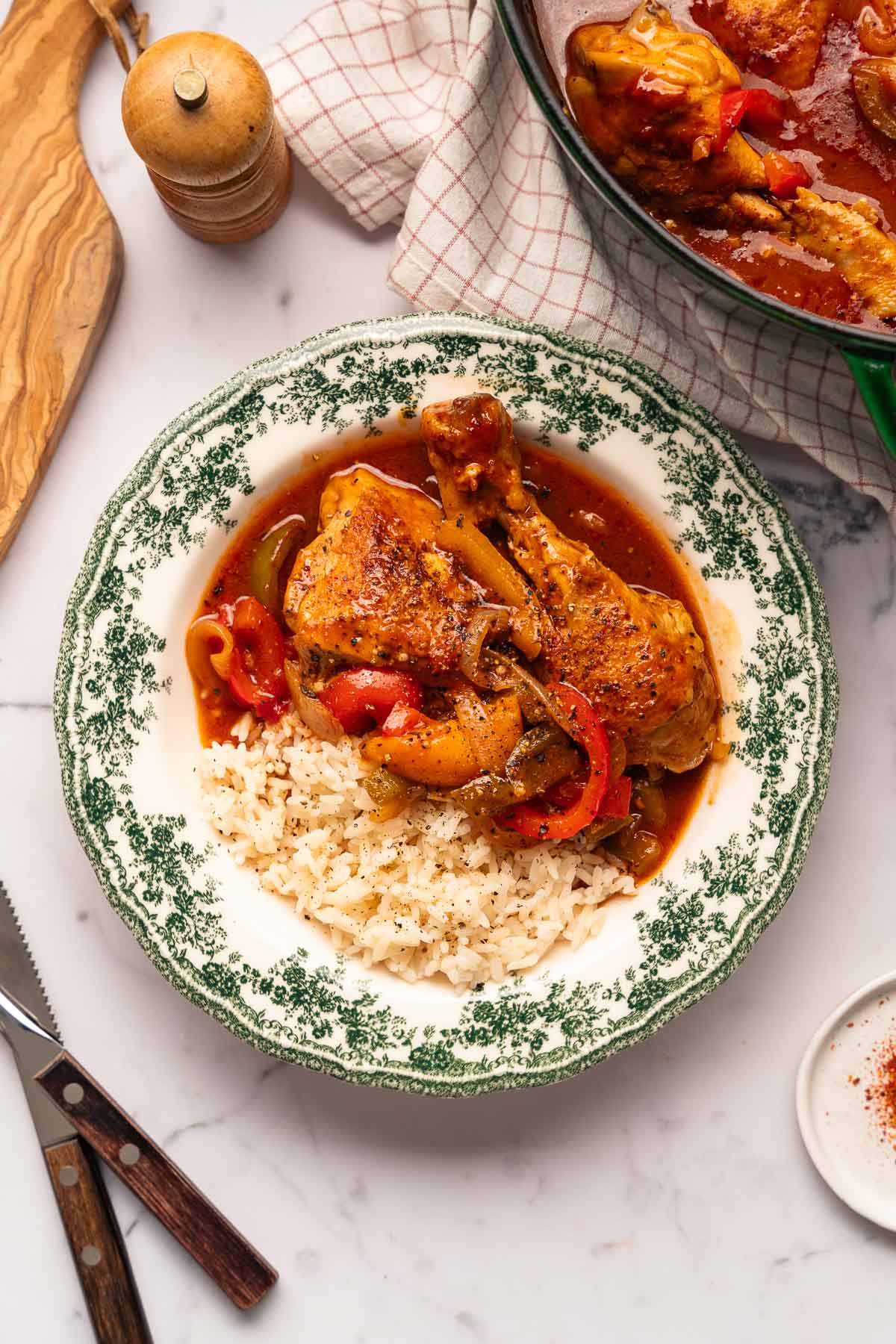 Une assiette de riz et de poulet basquaise avec des poivrons. Une casserole contenant le même plat est partiellement visible à l'arrière-plan, ainsi qu'une planche à découper en bois, un couteau et un moulin à poivre.