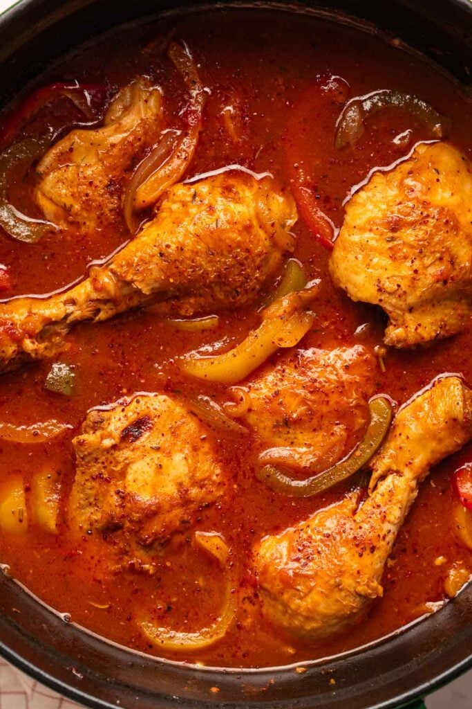 Close-up of a pot containing Basque chicken drumsticks simmered in a thick red sauce with sliced peppers.