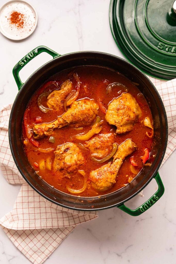 Ein grüner Topf mit Hühnchen in Tomatensoße mit Zwiebeln und Paprika, auf einem weiß karierten Küchentuch.