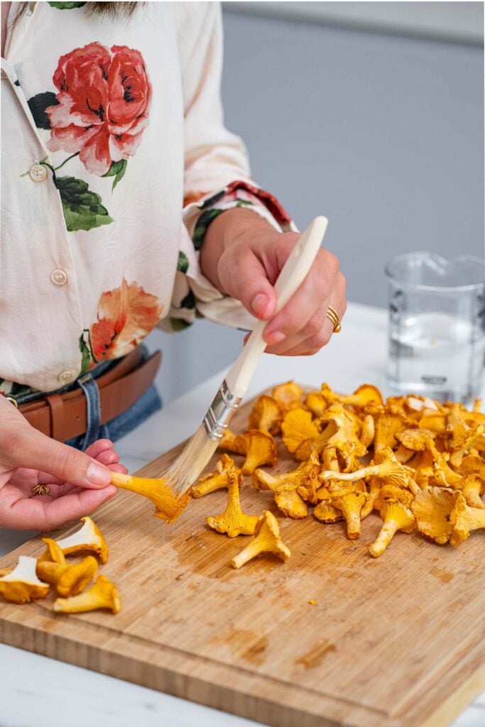 Une personne utilise une brosse pour nettoyer les champignons sur une planche à découper en bois, portant une chemise à imprimé floral et un jean.