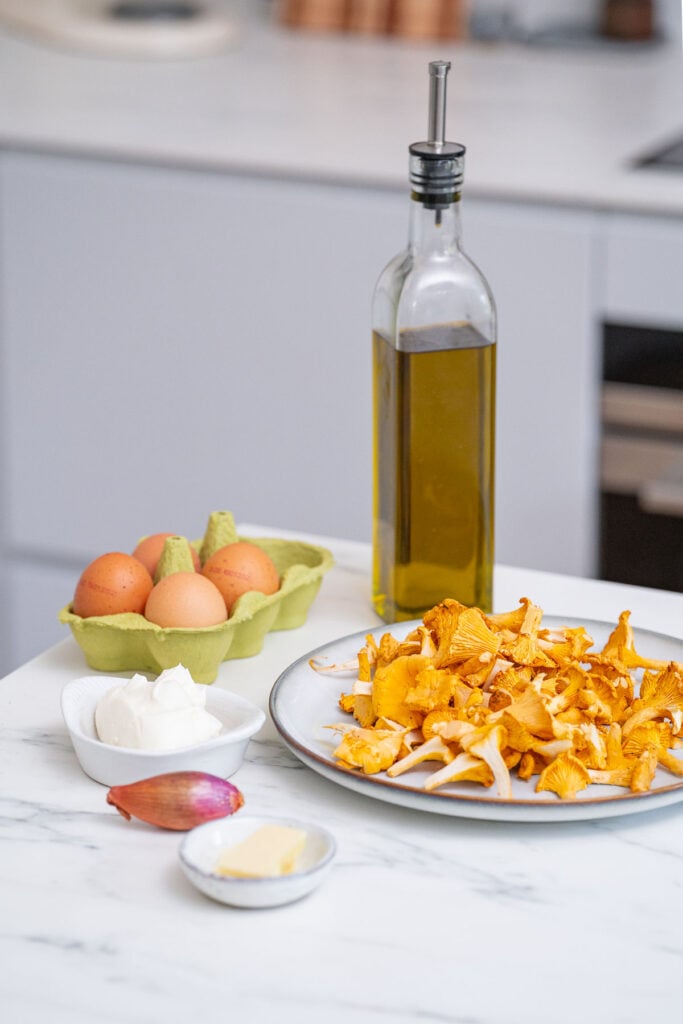 Un comptoir de cuisine avec des girolles sur une assiette, à côté d'une bouteille d'huile d'olive, des œufs dans un carton, une échalote, un petit bol de fromage à la crème et une noisette de beurre.