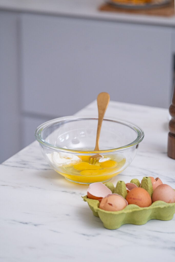 A glass bowl with broken eggs and a fork on a kitchen counter next to an open egg carton with broken eggshells.