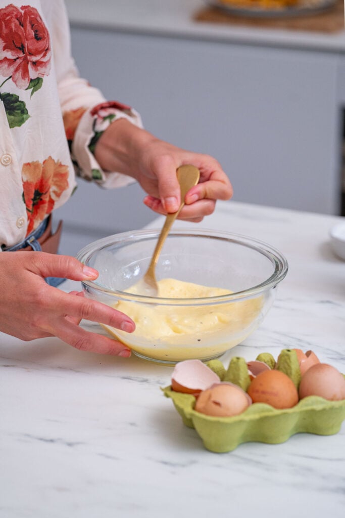 Person wearing a floral shirt whisking eggs in a glass bowl next to a carton of eggs on a marble countertop.