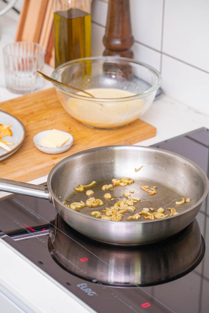 Une poêle en acier inoxydable avec des oignons sautés sur une cuisinière à induction. À l'arrière-plan, un bol en verre avec un mélange jaune, un petit plat avec du beurre et une bouteille d'huile sont posés sur une planche à découper.