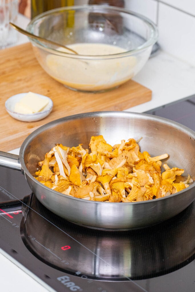 Une poêle en acier inoxydable avec des girolles en train de cuire sur une cuisinière à induction. Un bol de pâte et un plat de beurre sont posés sur une planche à découper en bois à l'arrière-plan.