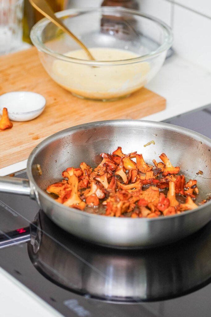 Une poêle en acier inoxydable avec des champignons sautés sur une cuisinière, avec une planche à découper, un petit bol et un autre bol contenant un mélange visible en arrière-plan.