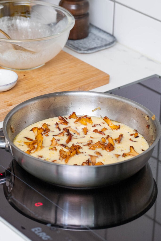 Une poêle sur une cuisinière contient une sauce crémeuse avec des tranches de champignons dorés. Un bol en verre et une planche à découper en bois sont visibles à l'arrière-plan.