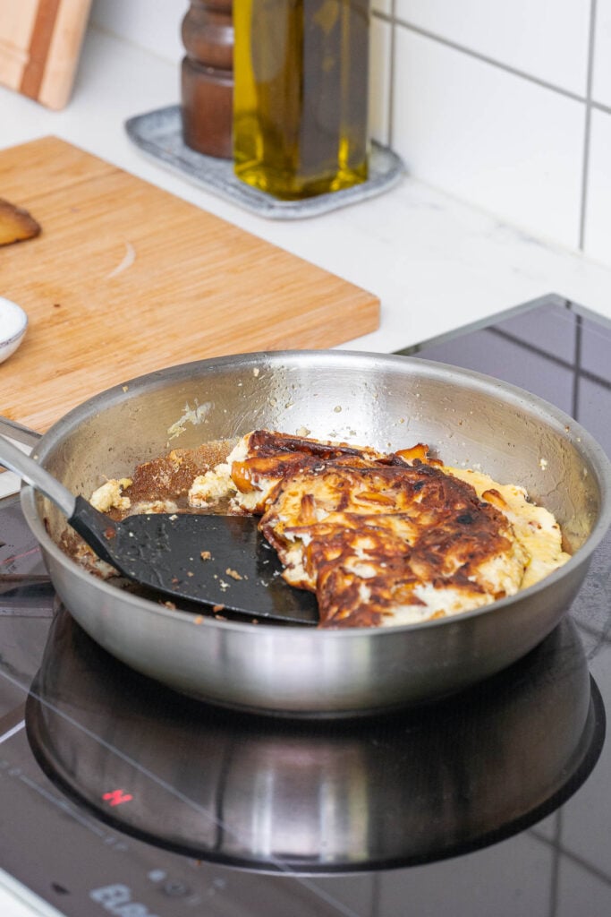 Une omelette brûlée dans une poêle en acier inoxydable sur une cuisinière à induction. Une spatule noire soulève partiellement l'omelette. Une planche à découper et une bouteille d'huile sont visibles en arrière-plan.