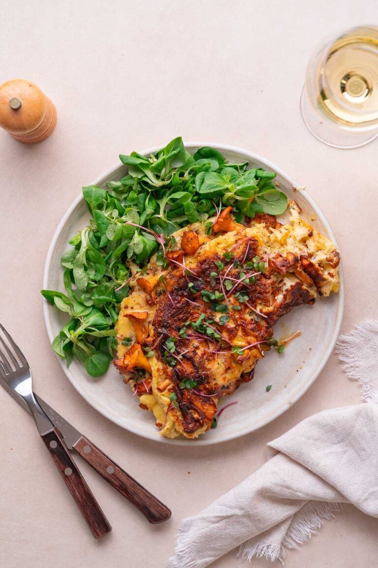 Une assiette de nourriture comprenant une omelette aux légumes, accompagnée d'une salade verte, d'un verre de vin blanc et d'ustensiles à côté.
Un moulin à poivre est également visible sur la table. 