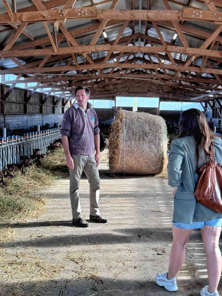 Two people stand chatting in a barn with a large bale of hay in the background.