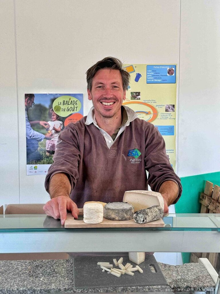 Un homme se tient debout derrière un comptoir présentant divers fromages, souriant. Il porte des vêtements décontractés et se tient devant des affiches sur un mur.