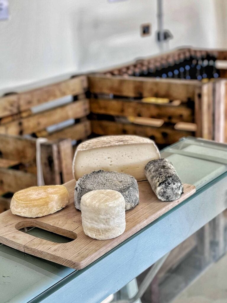 A wooden board presents four types of cheese, placed in front of wooden crates filled with bottles in the background.