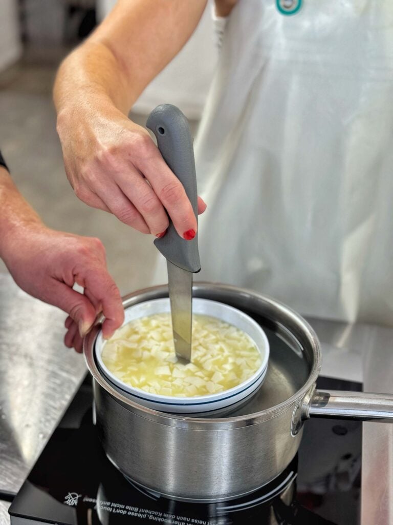 Jemand hält ein Messer senkrecht in eine Schüssel mit Paneer, die in einem mit Wasser gefüllten Topf steht, wahrscheinlich um die Konsistenz oder die Temperatur des Paneers zu überprüfen.