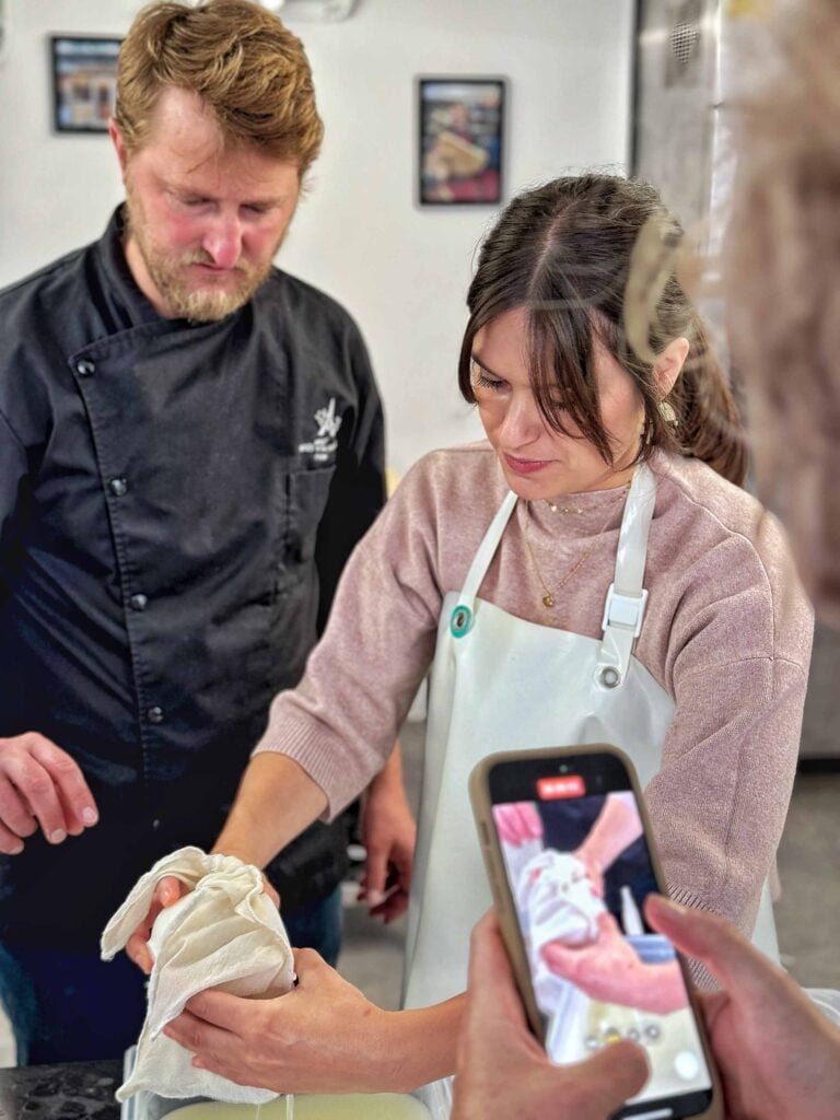 Une femme et un chef cuisinier se concentrent sur une tâche culinaire, la femme tenant un torchon blanc. Une personne au premier plan filme la scène avec un smartphone.