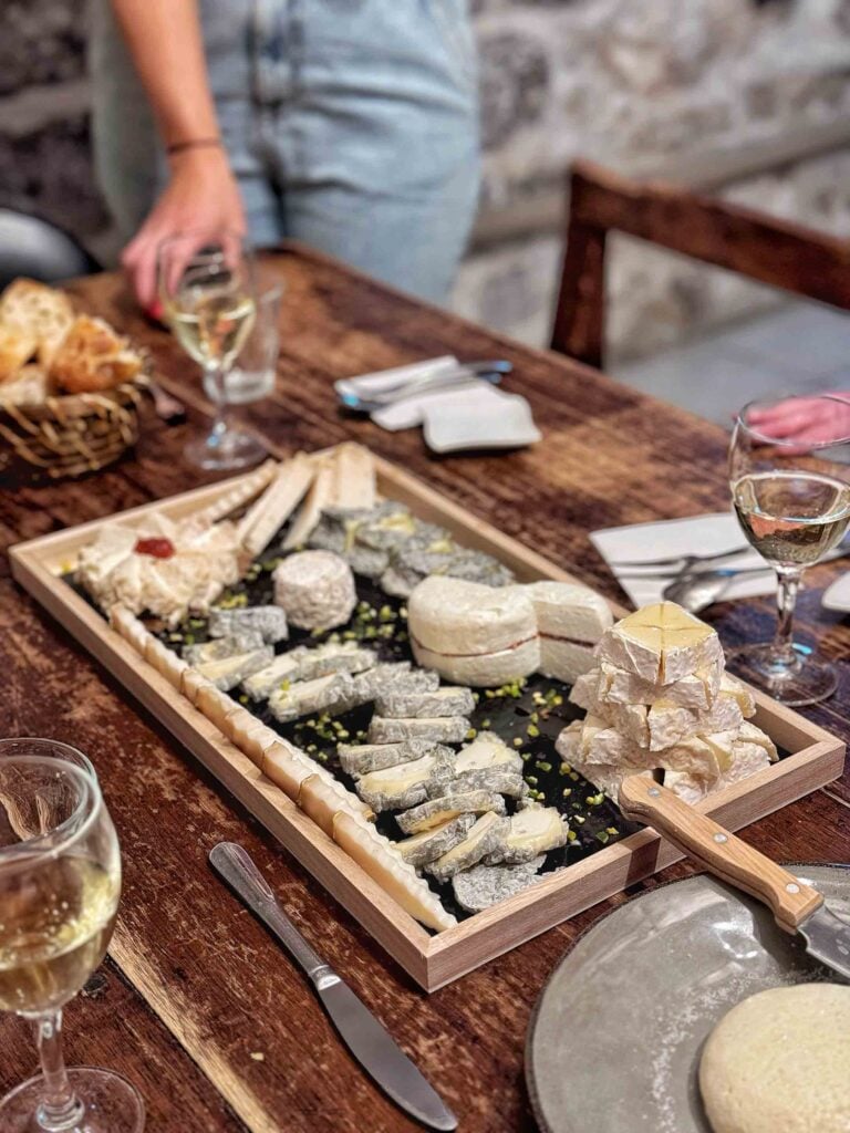 Une table en bois dressée avec divers fromages sur un plateau rectangulaire, des verres à vin, une corbeille de pain, des ustensiles et des serviettes. Une personne se tient debout près de la table.