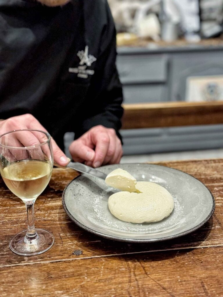 Person cutting a round cheese on a plate, with a glass of white wine next to her on a wooden table.