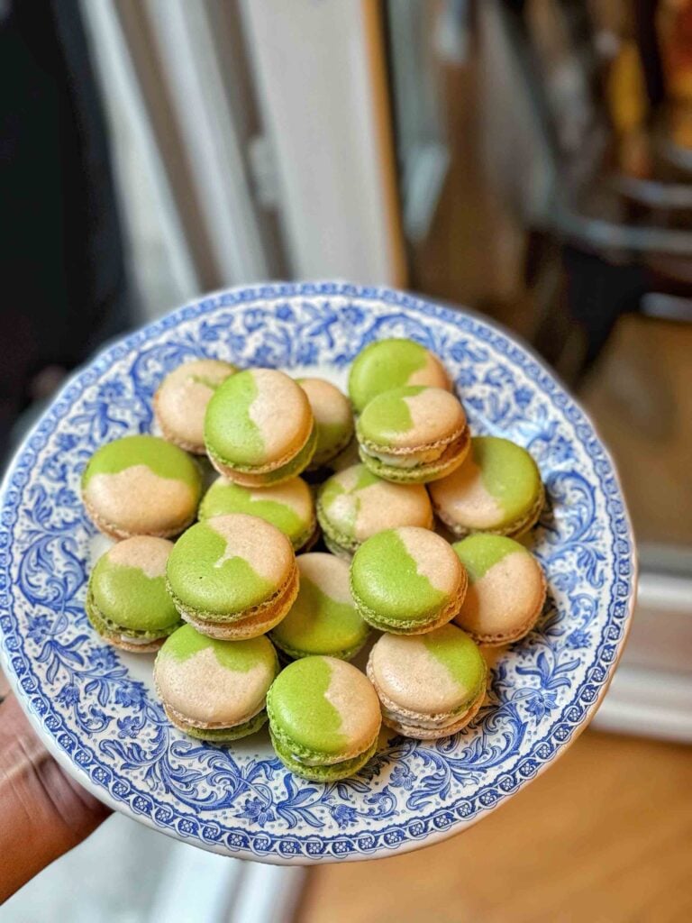 Une assiette décorative bleue et blanche accueille une pile de macarons verts et beiges. L'environnement évoque un décor intérieur.