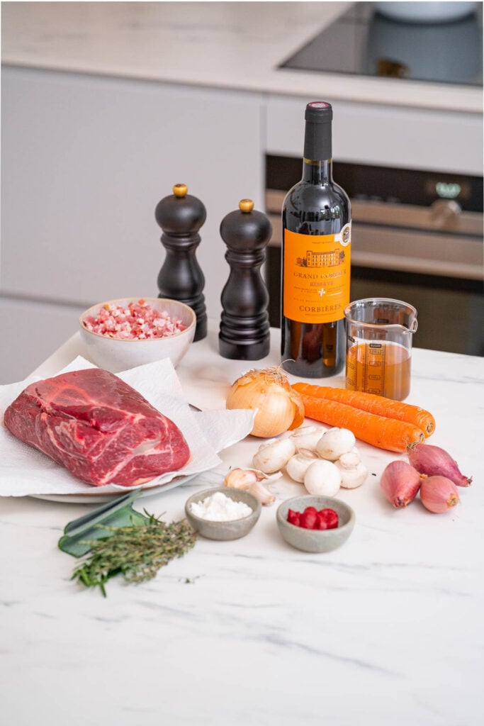 Kitchen ingredients displayed on a kitchen counter include a bottle of wine, raw beef, pepper mills, vegetables, herbs and small bowls containing flour and berries.