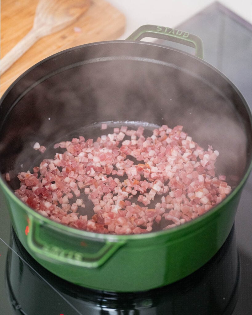 Gehackter Speck wird in einem grünen Topf auf dem Herd gekocht. Dampf entweicht aus dem Speck. Im Hintergrund ist ein Holzlöffel zu sehen.  