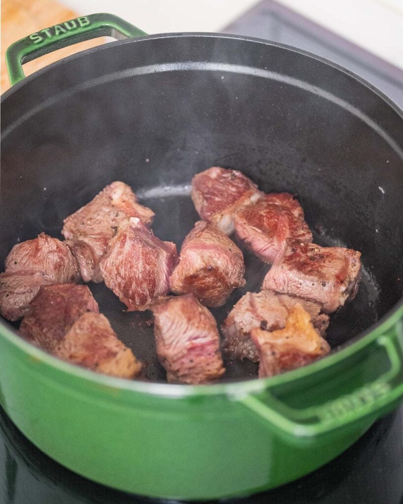 Pieces of meat searing in a green Staub pot on a stovetop.