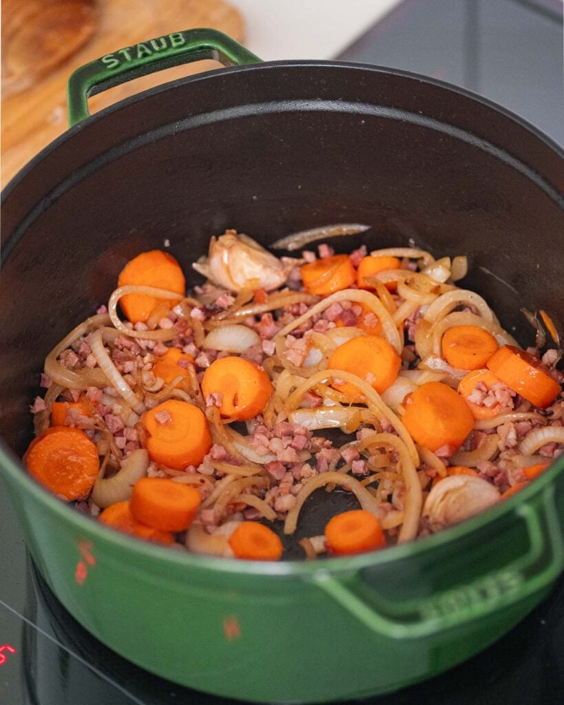 Grüner Topf auf dem Herd mit geschnittenen Karotten, Zwiebeln und Speckstücken.