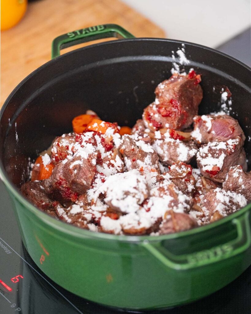 Chunks of raw meat with diced carrots and a little flour in a green saucepan on a stovetop.