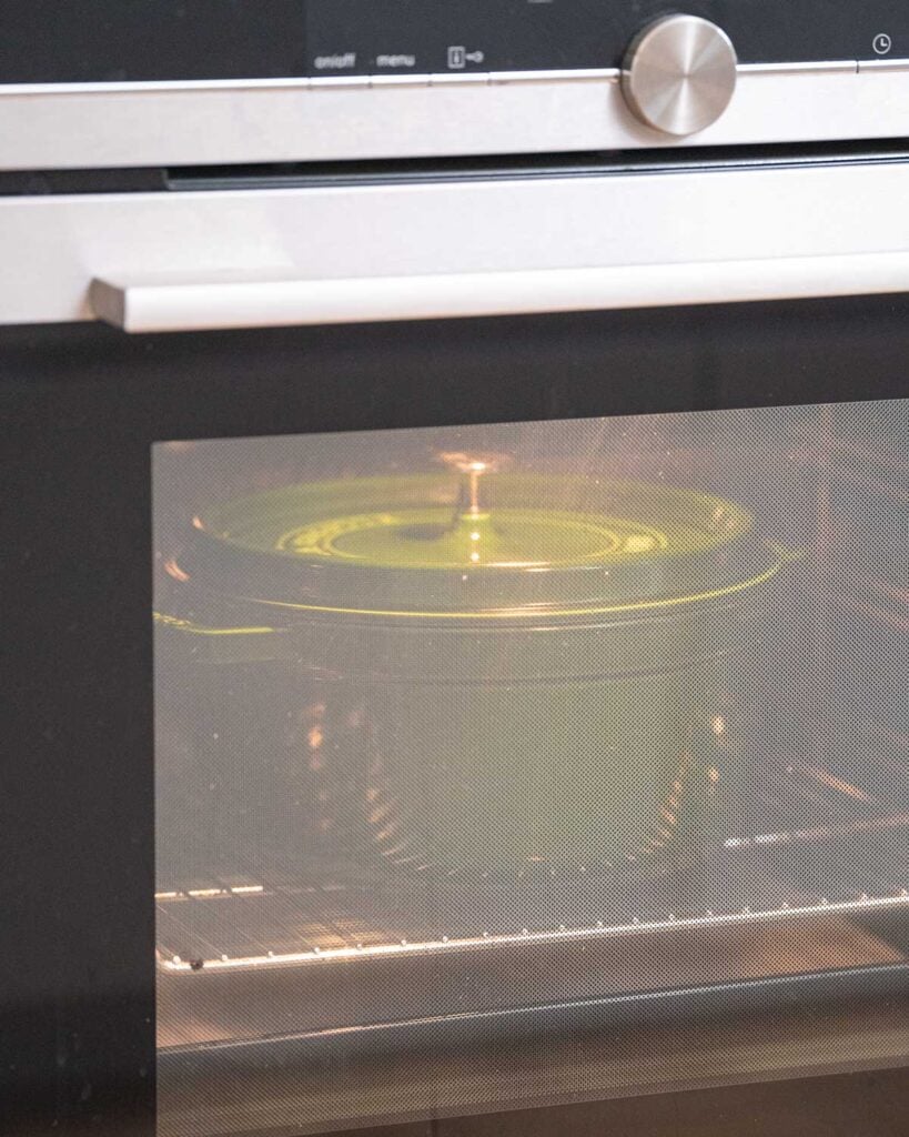 Une casserole verte avec un couvercle est visible sur une grille à l'intérieur d'un four à travers la porte du four.