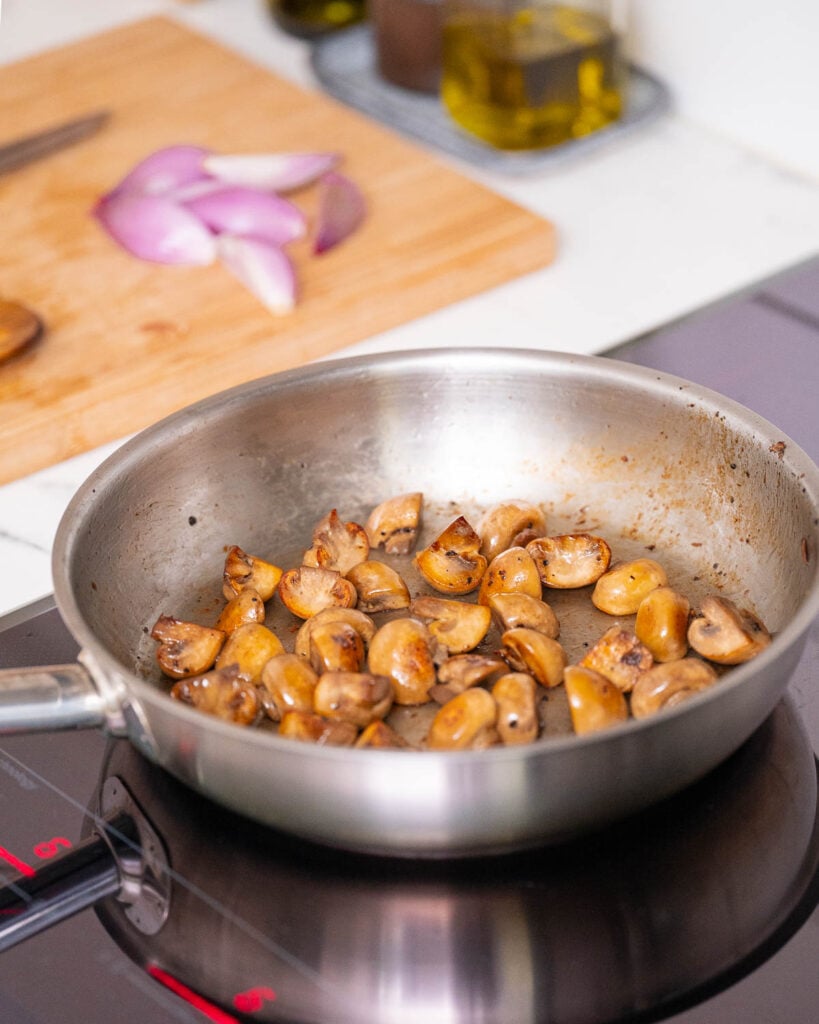 A pan with sautéed mushrooms on a stovetop; sliced onions on a cutting board in the background.