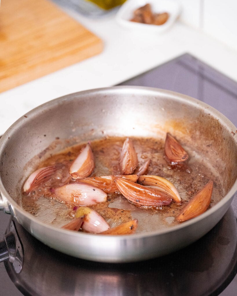 A stainless steel frying pan with caramelized shallots cooking on a stovetop. A wooden cutting board and a small dish are visible in the background. 