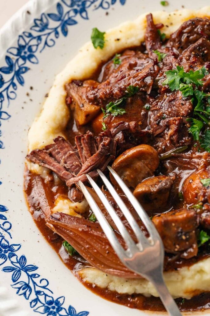 Une assiette de bœuf bourguignon avec du bœuf braisé et des champignons dans une sauce riche, servie avec une purée de pommes de terre crémeuse. Une fourchette est placée sur le bord du plat.