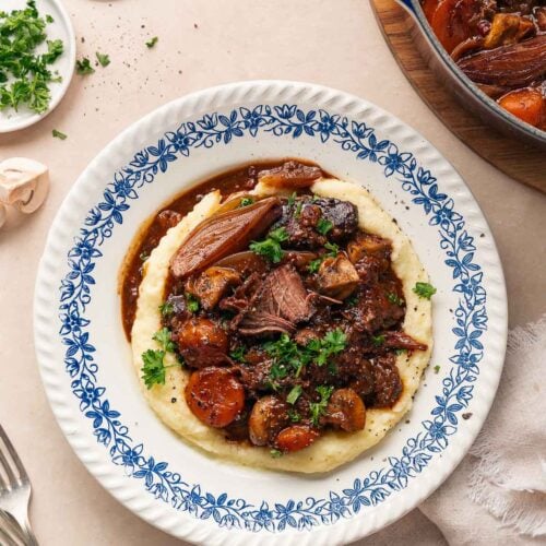 A plate of beef bourguignon served over mashed potatoes, garnished with parsley. A pot of this rich stew, a napkin and forks are nearby.