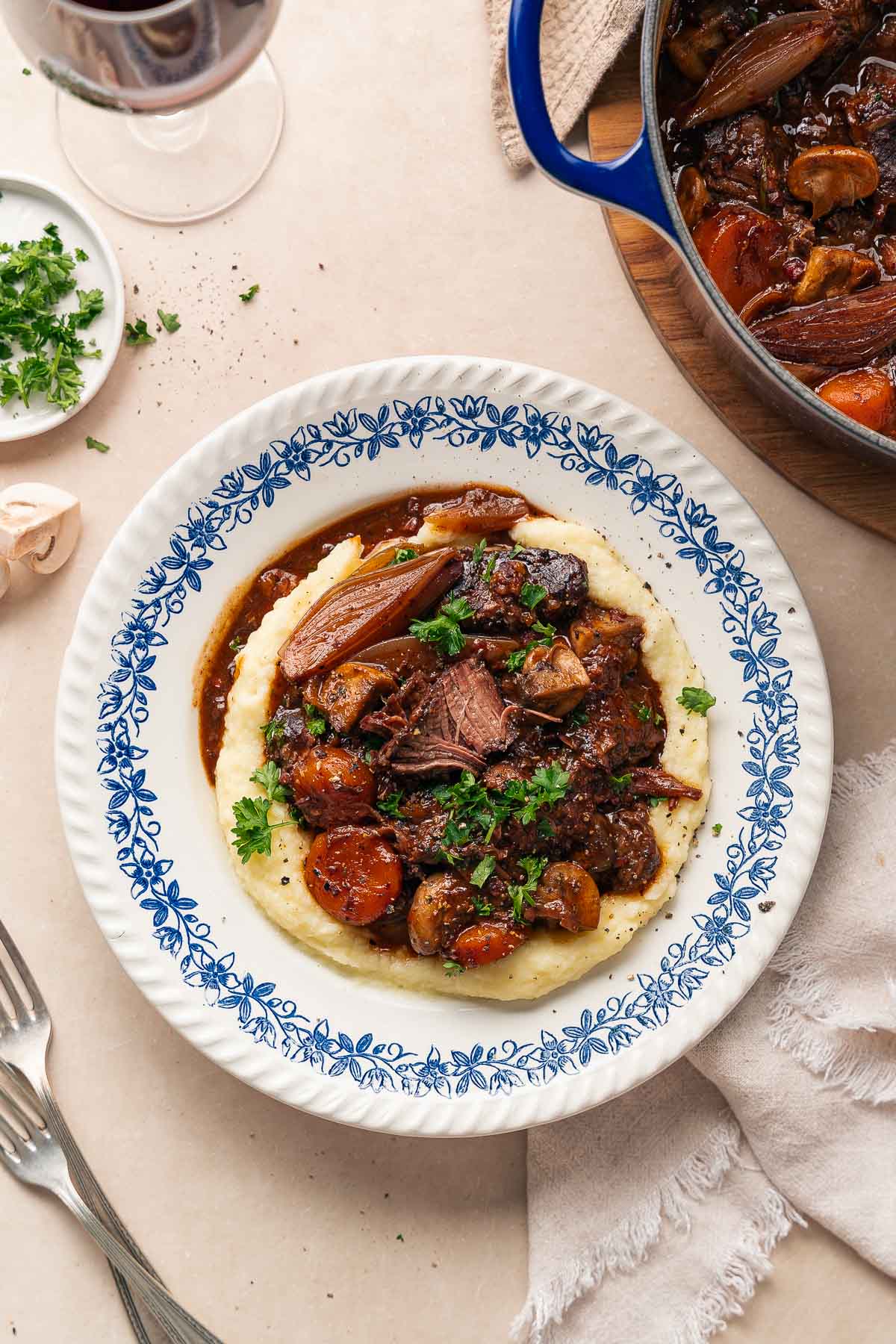 A plate of beef bourguignon served over mashed potatoes, garnished with parsley. A pot of this rich stew, a napkin and forks are nearby. 