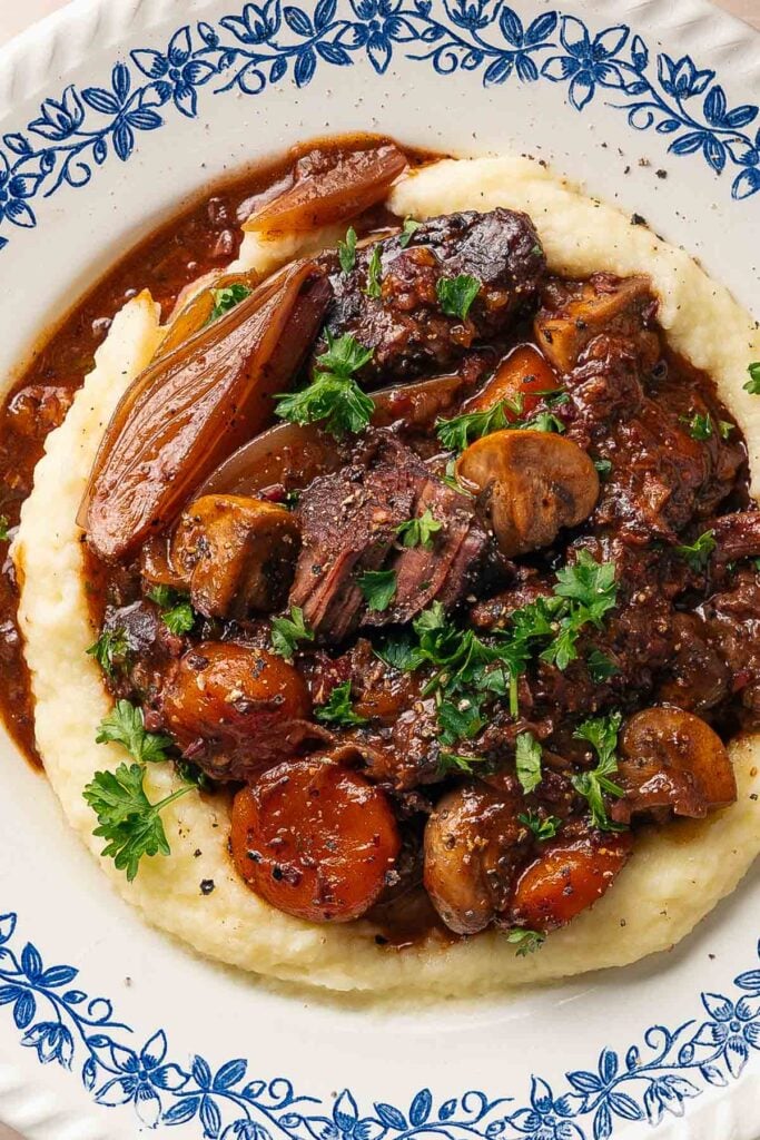 Un bœuf bourguignon copieux avec du bœuf tendre, des carottes, des champignons et des herbes servi sur une purée de pommes de terre crémeuse dans une assiette décorative.