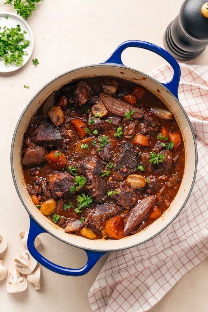 A blue pot filled with boeuf bourguignon, composed of tender pieces of beef, carrots, onions and herbs, rests on a checkered cloth.