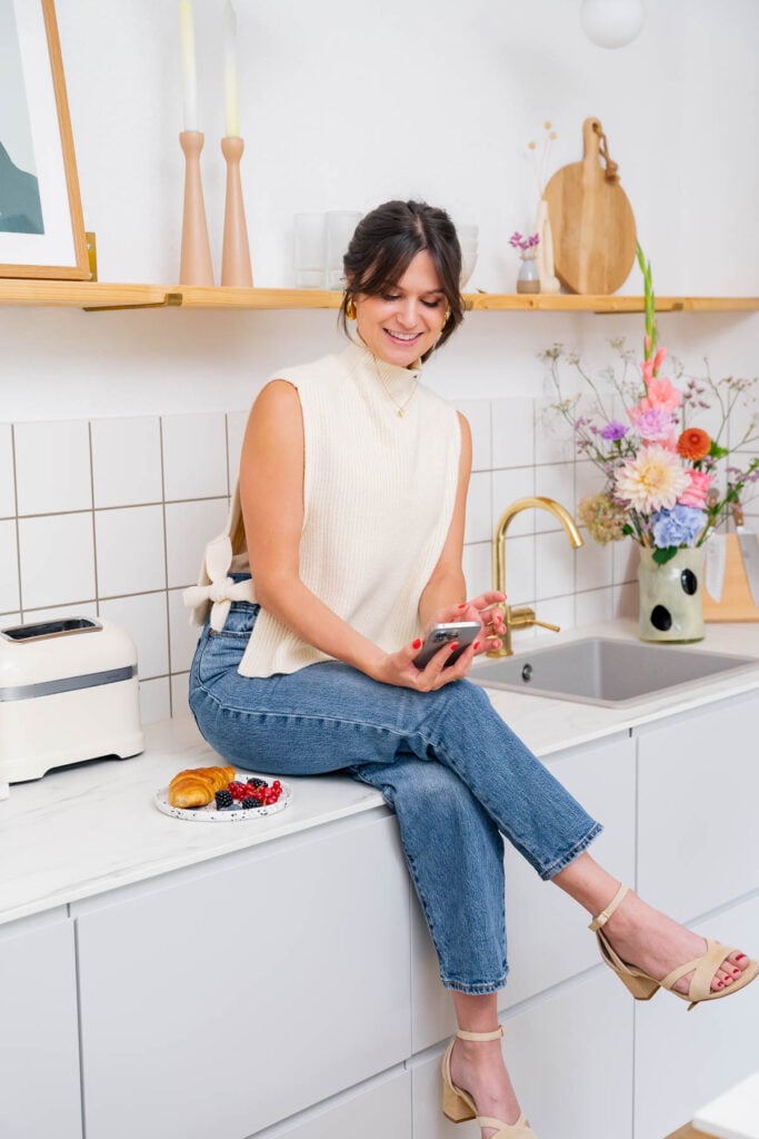 Géraldine Leverd est assise  sur le comptoir d'une cuisine. Elle porte un haut sans manches et un jean et a une main pensive sur le menton. Un croissant et un vase de fleurs sont posés à proximité, ce qui contribue à l'atmosphère paisible.