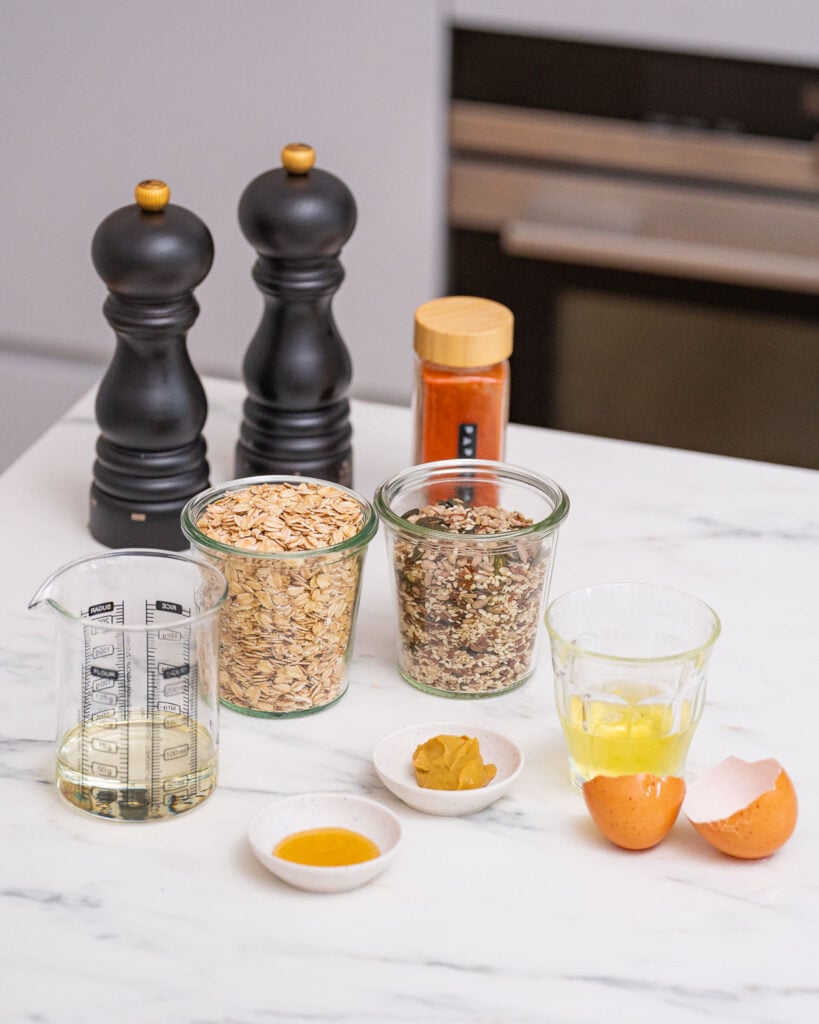 Ingredients on a kitchen counter include rolled oats, mixed seeds, honey, mustard, oil in a measuring cup, broken eggshells, and pepper mills in the background.