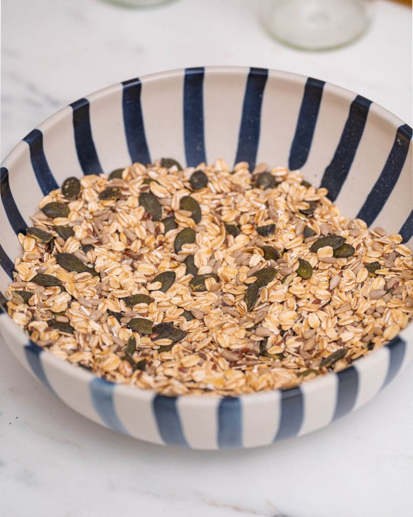 A blue and white striped bowl holds oats mixed with pumpkin seeds on a marble surface.